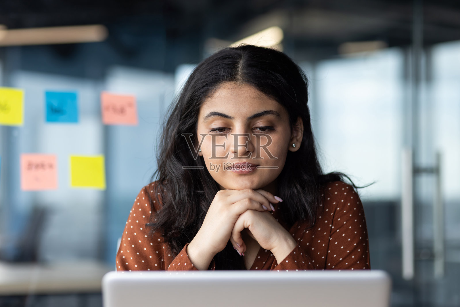 一位专注的年轻女商人在现代办公室里使用笔记本电脑工作。她有着深色头发，穿着棕色上衣，背景中有便签纸。照片摄影图片