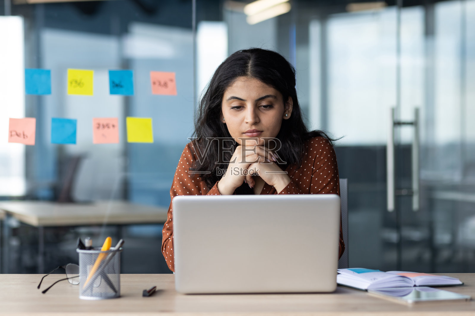 专注的年轻女性在现代办公室的笔记本电脑上工作，玻璃墙上贴满了五颜六色的便利贴。照片摄影图片