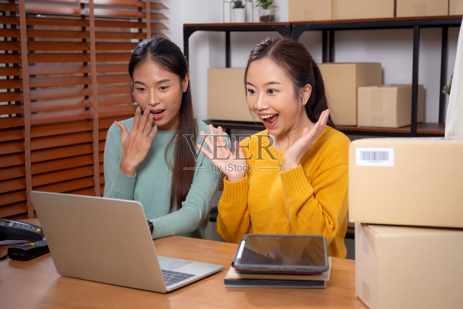 两位年轻的亚洲女性在办公室使用平板电脑和笔记本电脑查看商店评价，面带成功和愉快的笑容，展现出创业者或在线卖家的形象，涉及电子商务或初创企业，小型企业。照片摄影图片