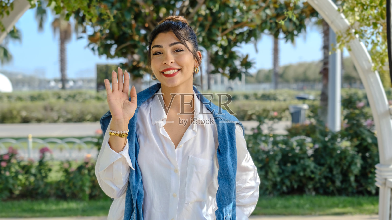 一位年轻的棕发女商人在春天的树木繁茂的公园里，面向镜头并挥手。照片摄影图片