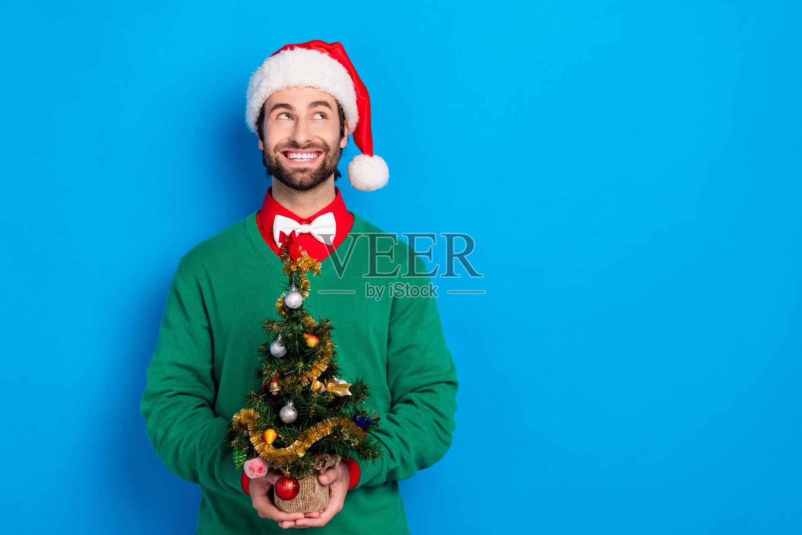 一位思考中的帅气男士双臂抱着一棵小新年树装饰，望向空旷的空间，背景为蓝色。照片摄影图片