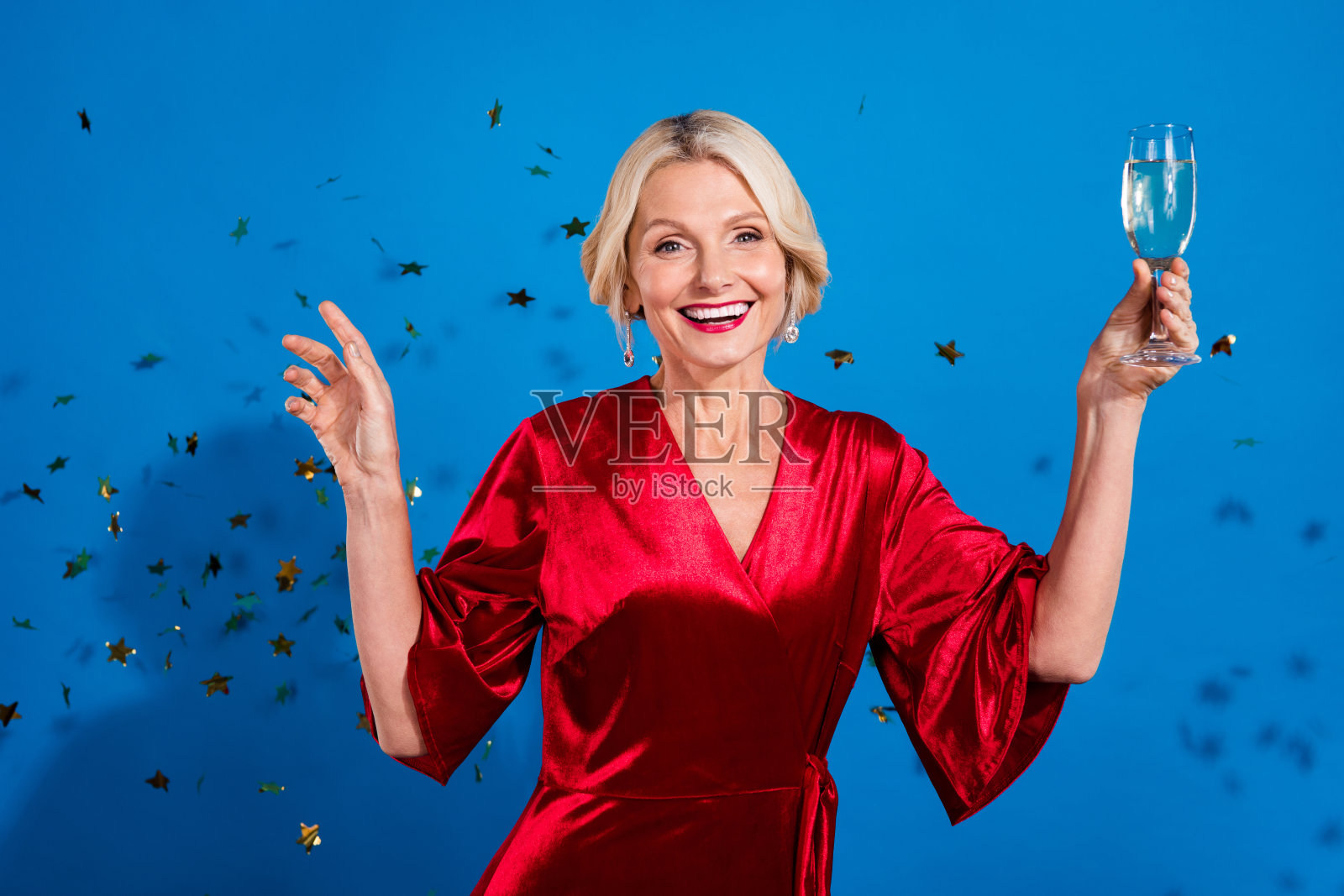 兴奋的时尚退休女士穿着闪亮的红色服装，举起酒杯享受圣诞派对，背景为蓝色。照片摄影图片