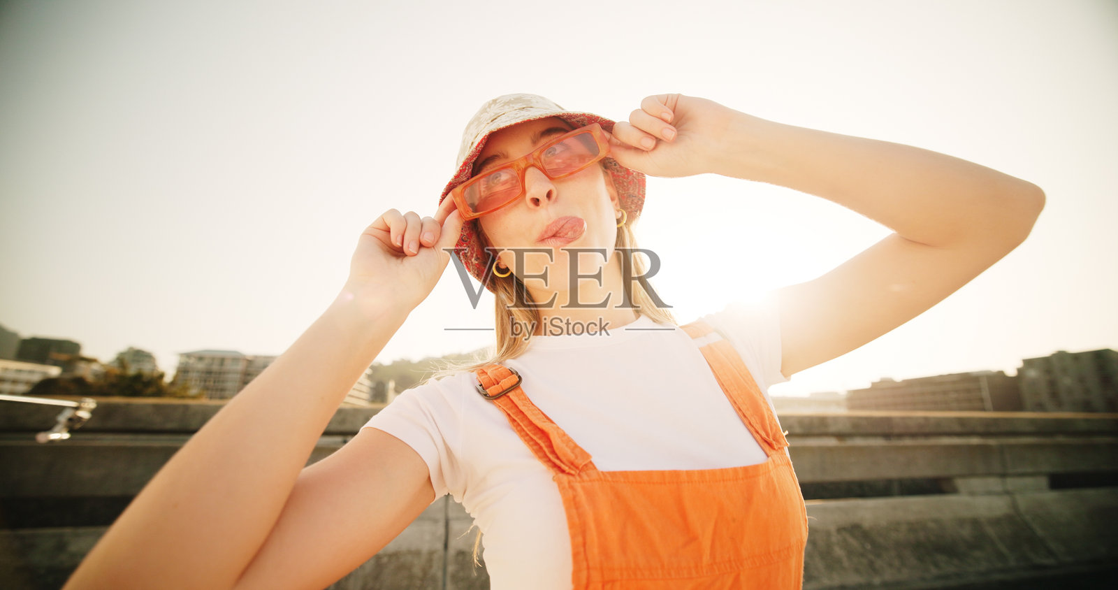 城市中戴着太阳镜的女人吐舌头，展现出时尚、顽皮和搞笑的表情，站在地铁桥上。女孩在阳光下戴着眼镜和帽子，开玩笑，表情搞怪，穿着时尚街头服饰。照片摄影图片