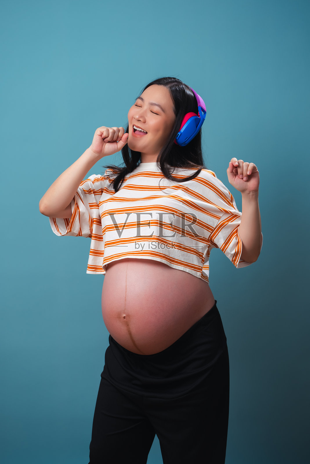 戴着耳机的亚洲孕妇在蓝色背景下独自享受舞蹈。照片摄影图片