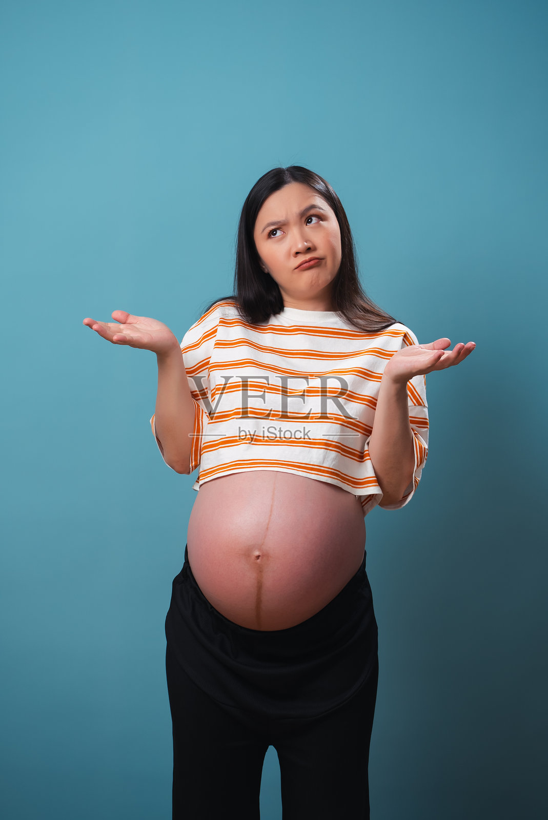 亚洲孕妇感到烦恼，耸耸肩，独自站在蓝色背景前。照片摄影图片