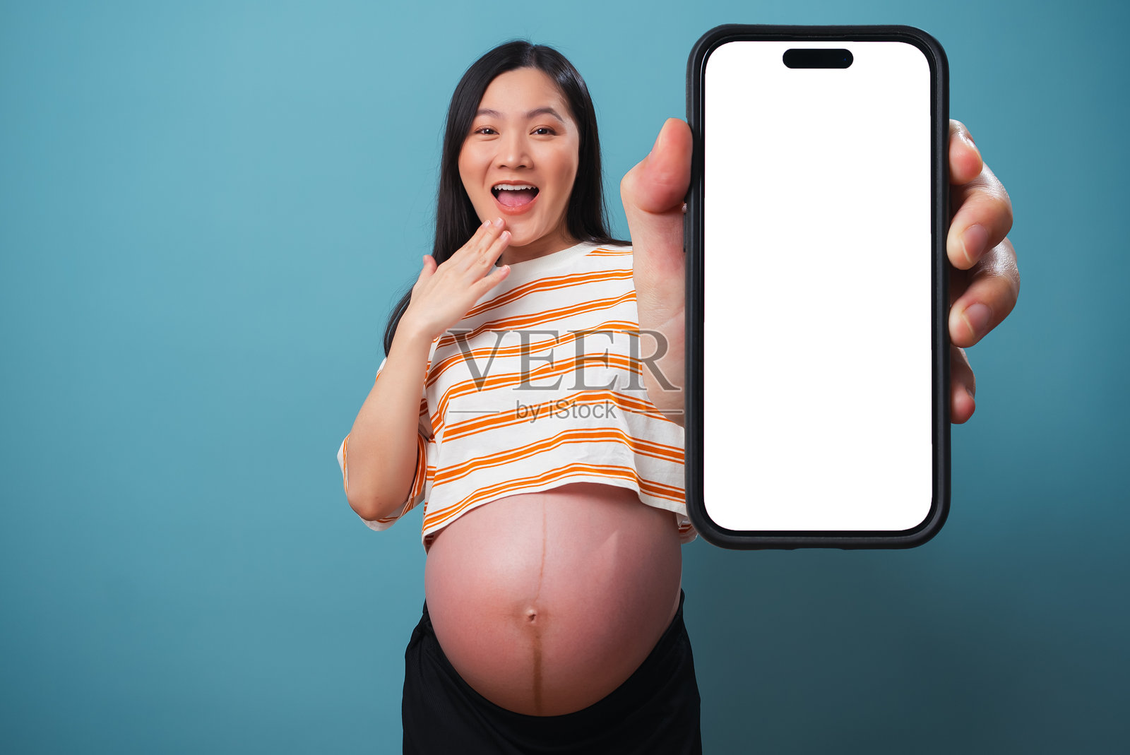 亚洲孕妇开心兴奋地展示智能手机上的白色屏幕，独自站在蓝色背景前。照片摄影图片