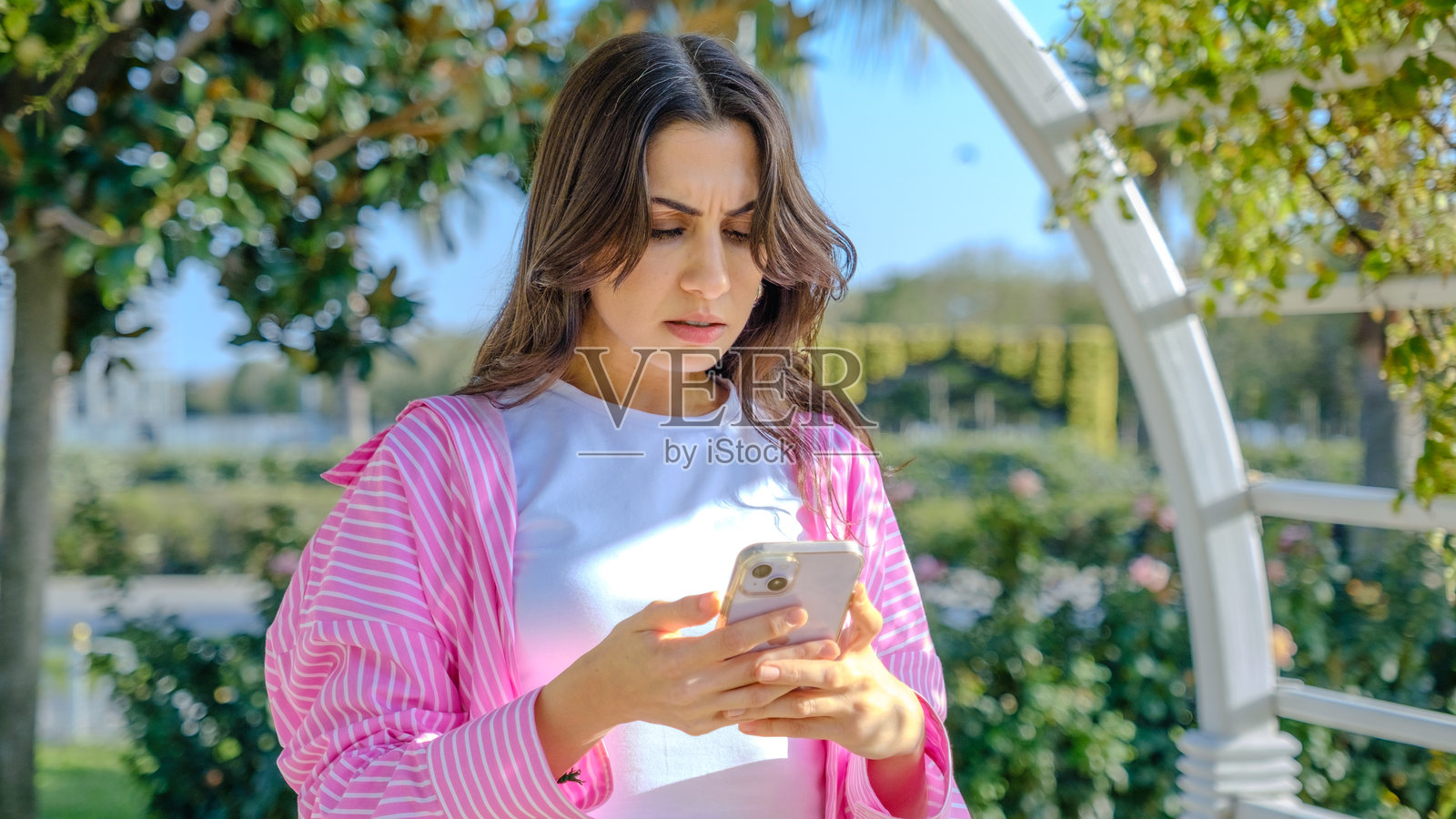 一位年轻的棕发女商人在春天的树木繁茂的公园里愉快地使用手机。照片摄影图片
