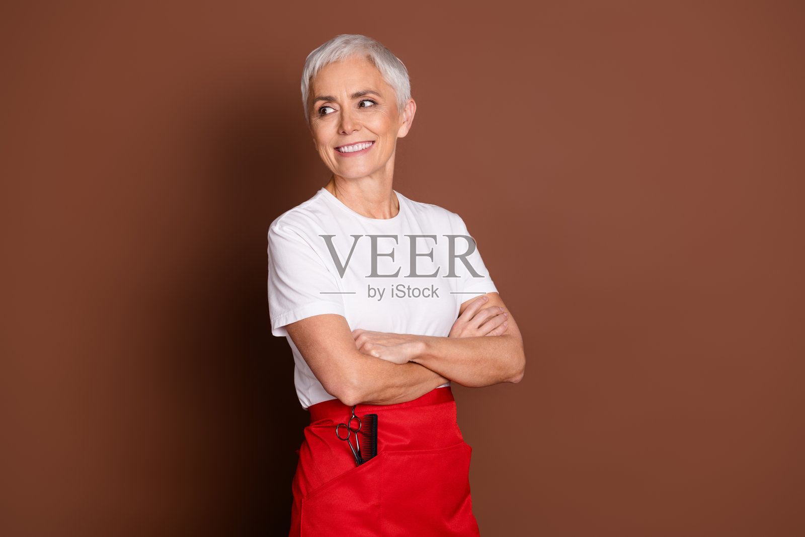 穿着制服的资深女性专业美发师，双手交叉，凝视空旷的空间，背景为棕色。照片摄影图片