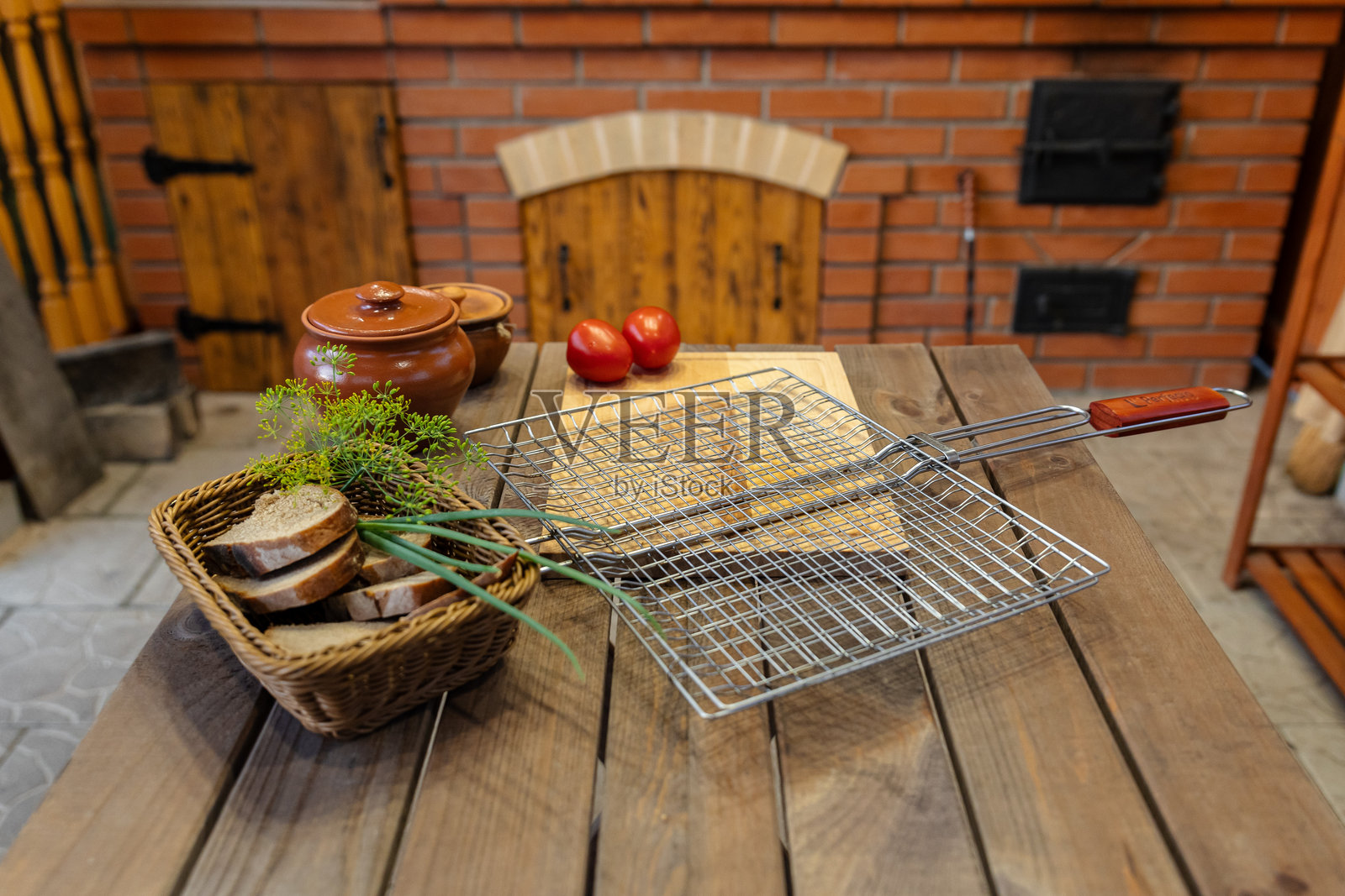 户外烧烤的餐桌布置。一个金属烤架放在木桌上，旁边有面包片、西红柿和陶瓷花盆。照片摄影图片