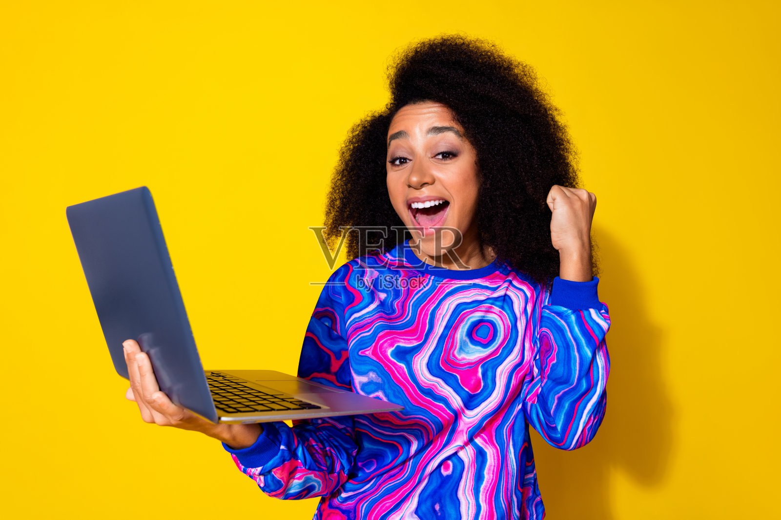 吸引人的年轻女性在黄色背景下穿着时尚的彩色蓝色衣服，手持笔记本电脑，展现胜利的姿态。照片摄影图片