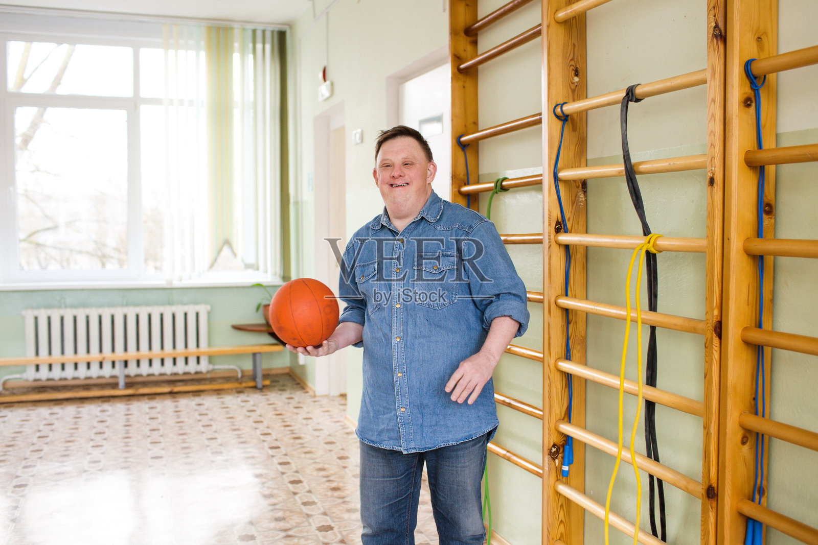 一位年轻的唐氏综合症男孩手握篮球，灿烂微笑，展现了他对运动的热爱和积极的态度。照片摄影图片