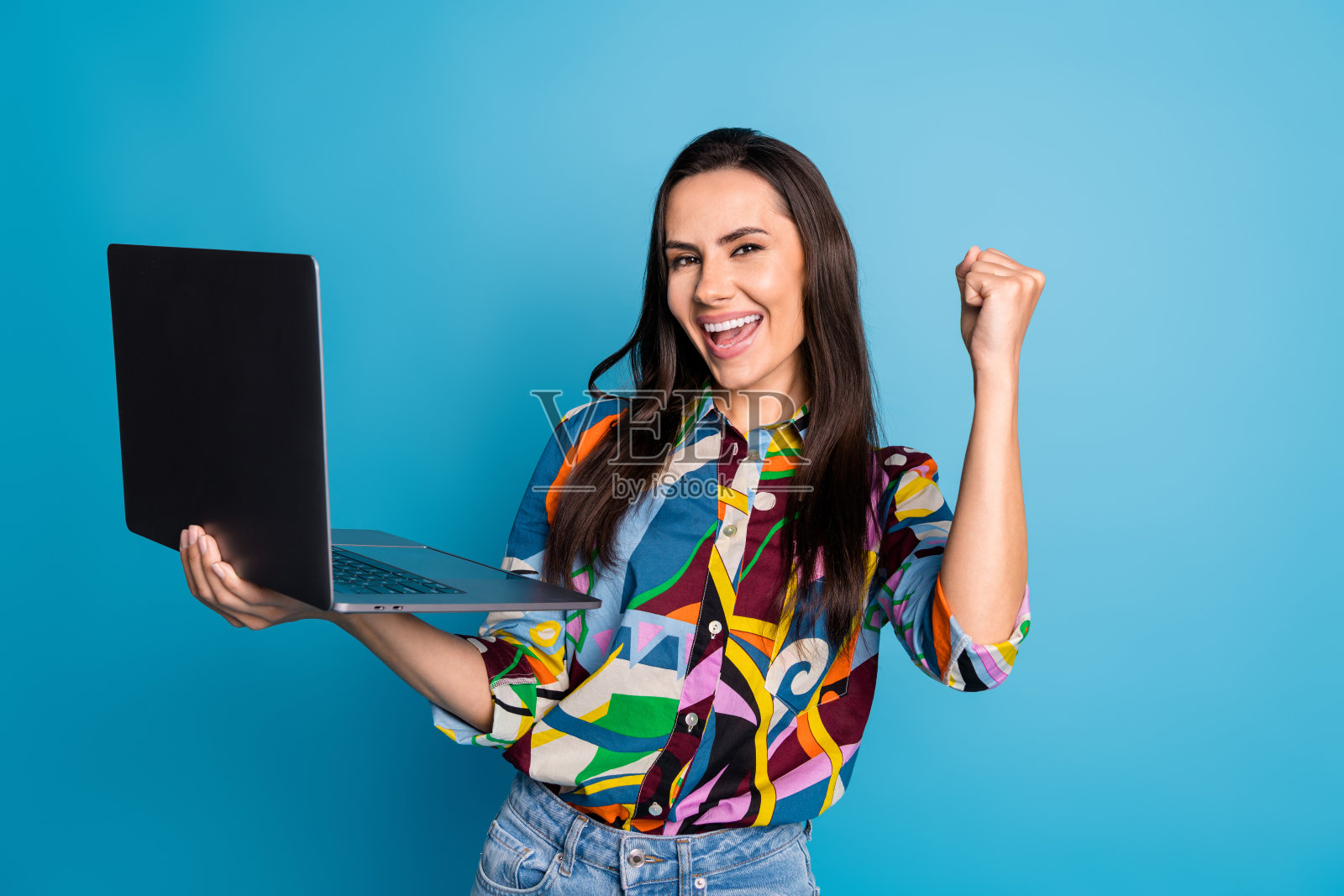 一位兴奋的女性，直发，穿着印花衬衫，手持笔记本电脑，因赌博获胜而大喊“是的”，背景为蓝色。照片摄影图片