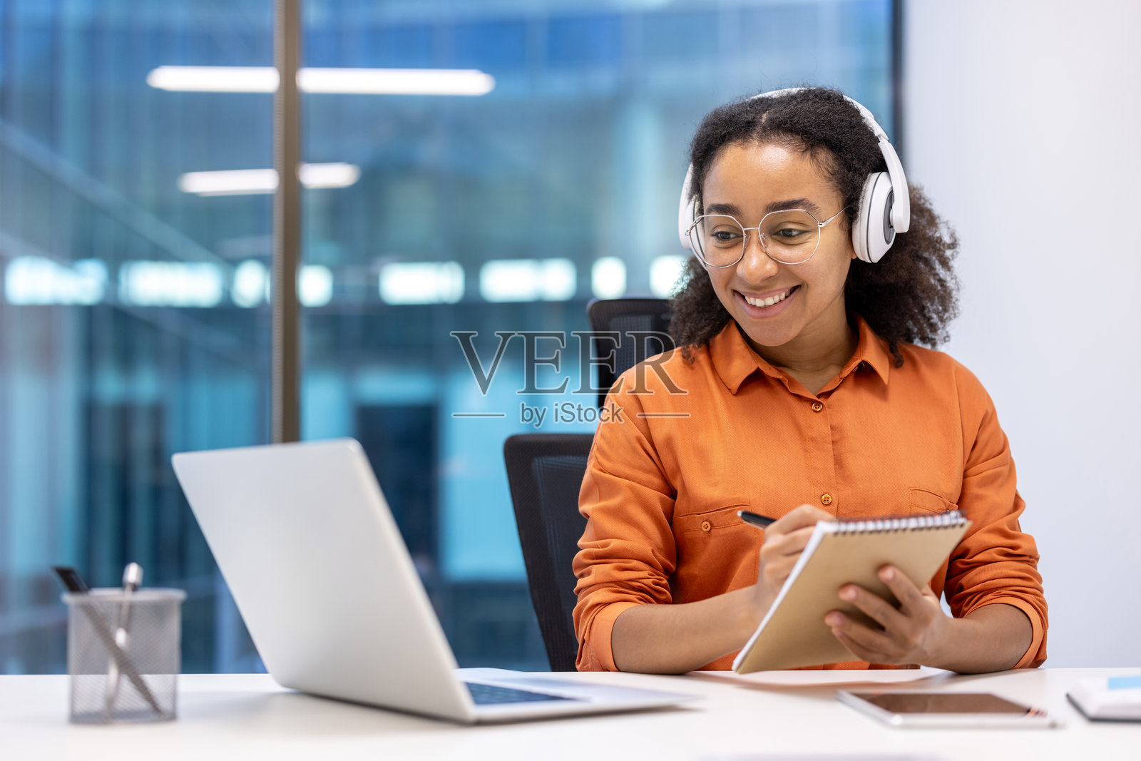 年轻女性在现代明亮的办公室里，戴着耳机，微笑着使用笔记本电脑和记事本，享受学习的乐趣。照片摄影图片