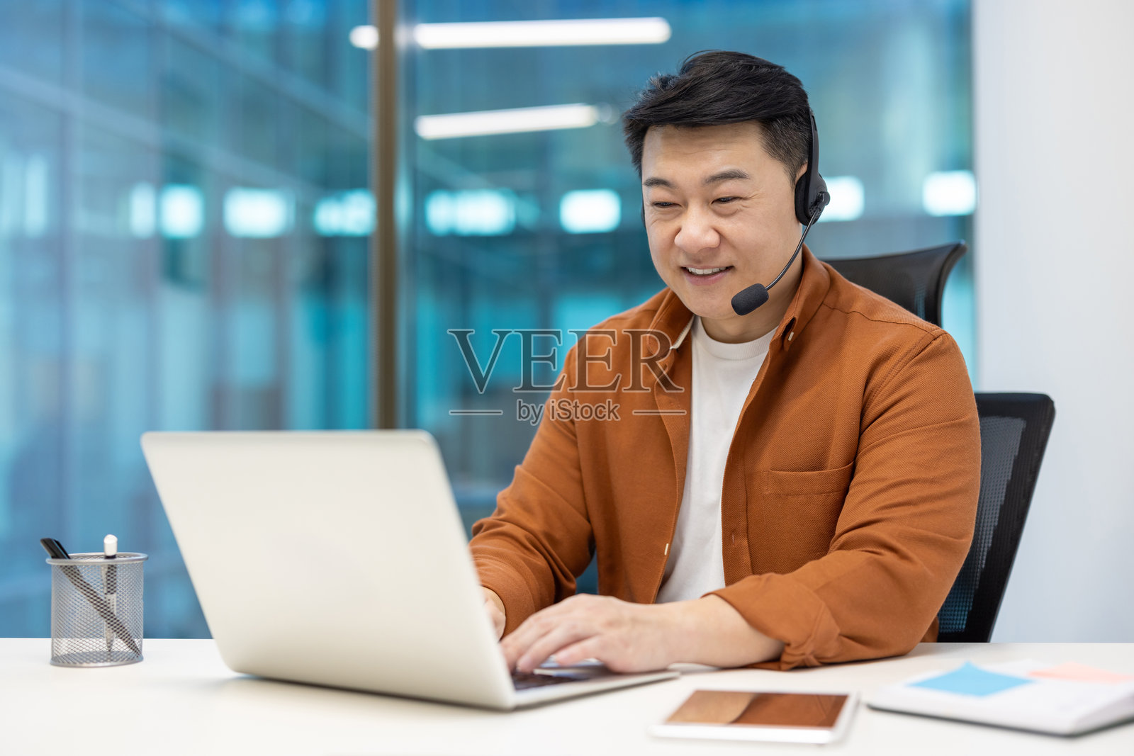 亚洲商人在现代办公室里戴着耳机，微笑着看着笔记本电脑，桌上放着智能手机，穿着休闲服，展现出高效的工作和沟通能力。照片摄影图片