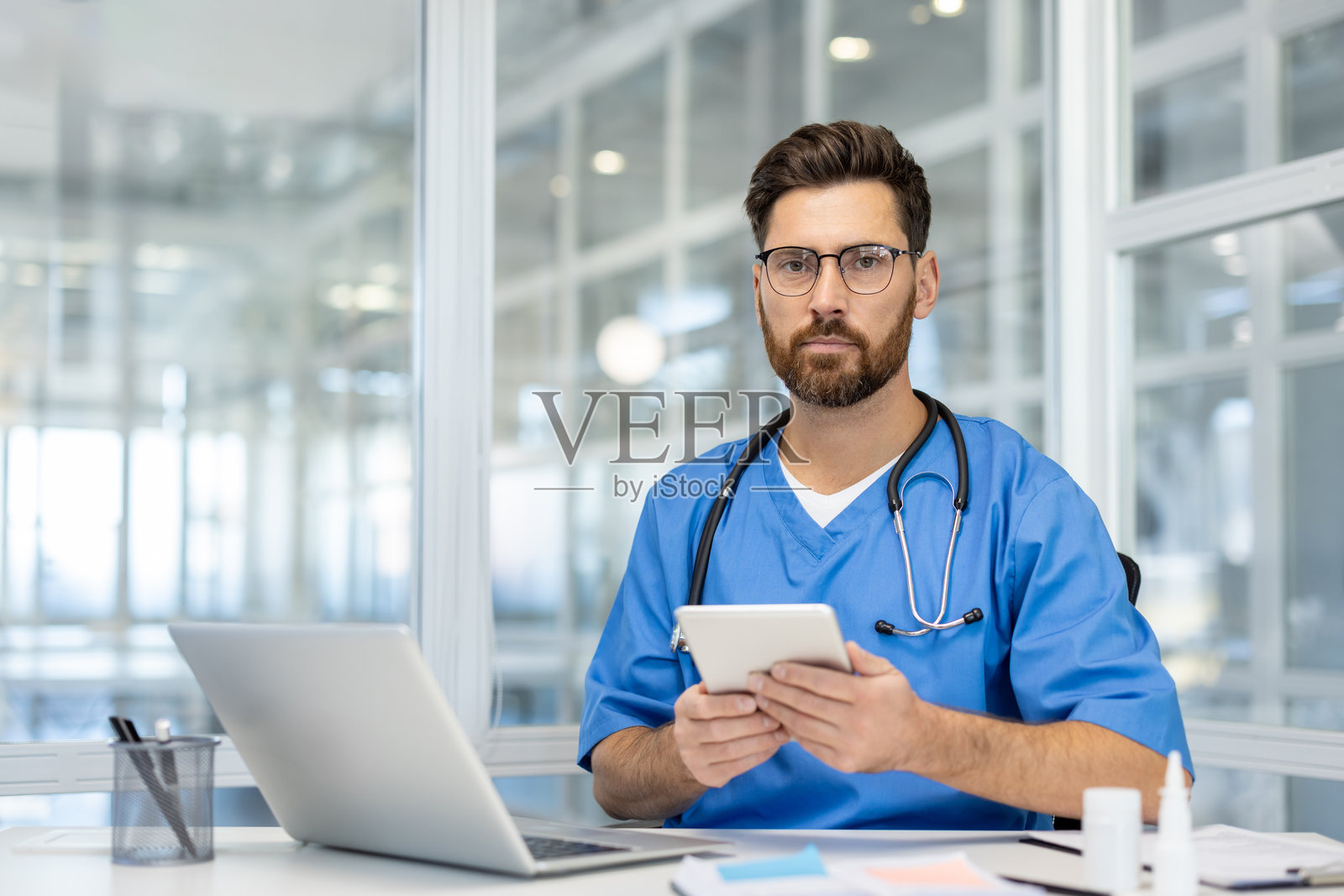 身穿蓝色手术服的专业男性医生在现代办公室环境中手持平板电脑，桌上有笔记本电脑和听诊器，传达医疗保健中的科技与专业精神。照片摄影图片