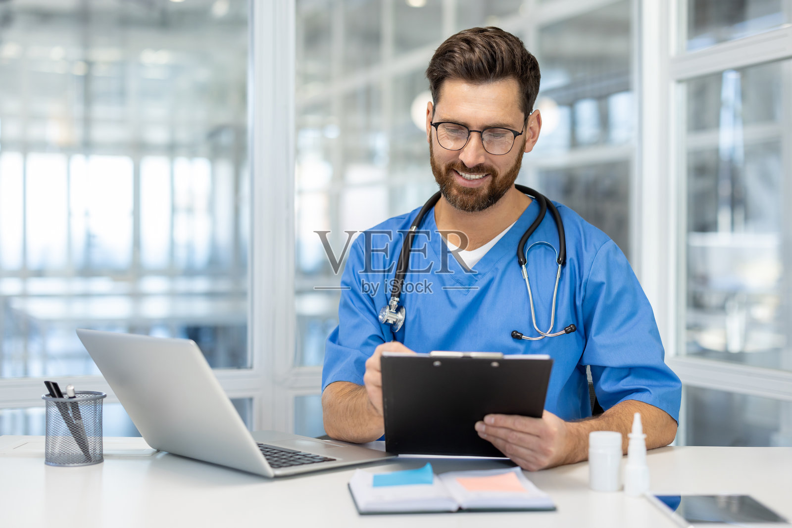 穿着蓝色医务工作服的医疗专业人士在现代办公室中使用笔记本电脑和夹子，自信地微笑着，适合医疗工作。照片摄影图片