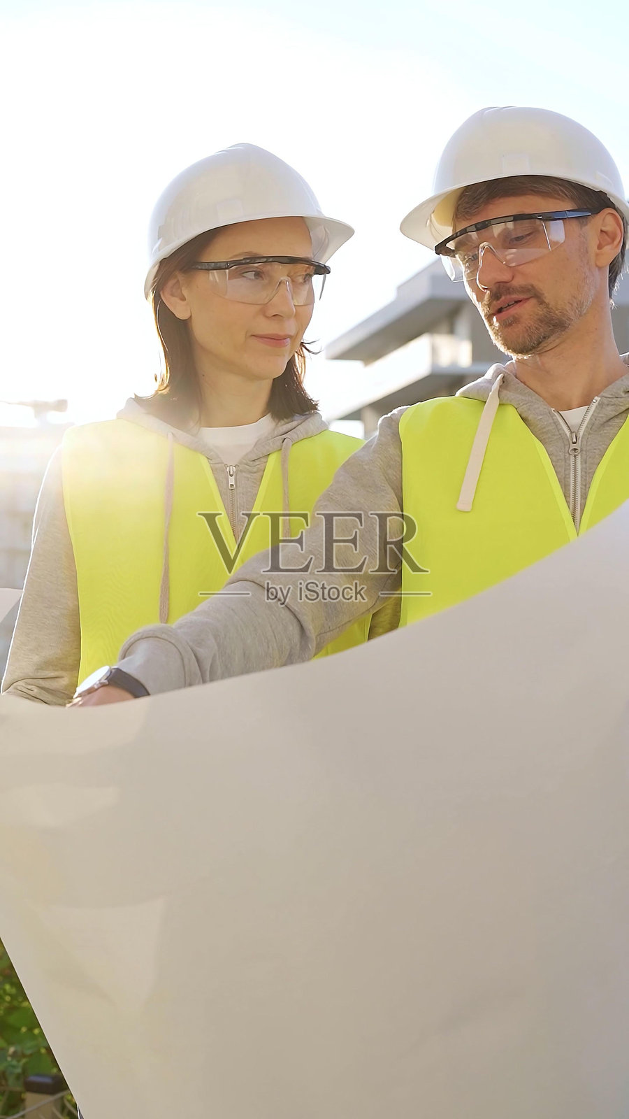 两位穿着安全帽和马甲的建筑师在日出时分的建筑工地上讨论蓝图，前方垂直视角。建筑工程与团队合作概念。照片摄影图片