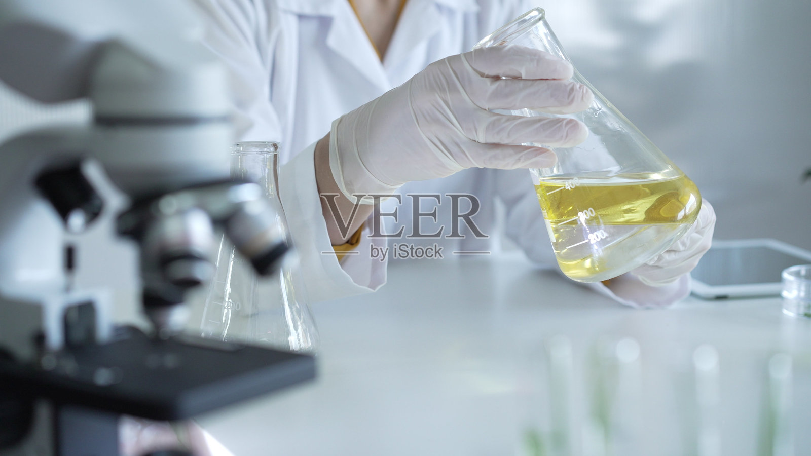 显微镜靠近一位穿着实验室外套和白色防护手套的科学家，他在实验室环境中手持装有黄色油状液体的烧杯，特写镜头。医学与科学概念。照片摄影图片