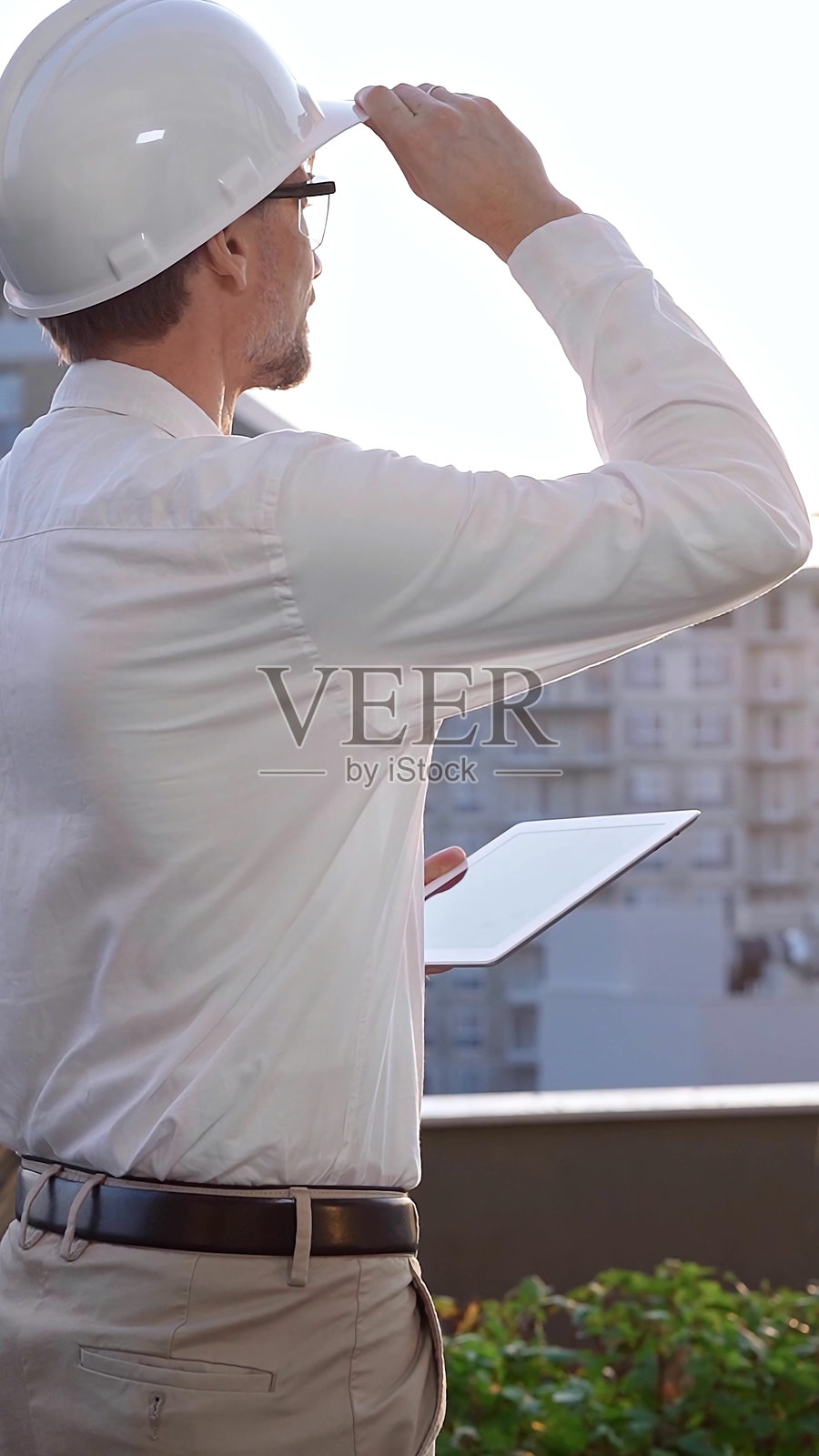 一名男性建筑工程师穿着白衬衫和安全帽，在日出时分检查建筑工地，使用数字平板电脑，背面和垂直视角。建筑概念照片摄影图片