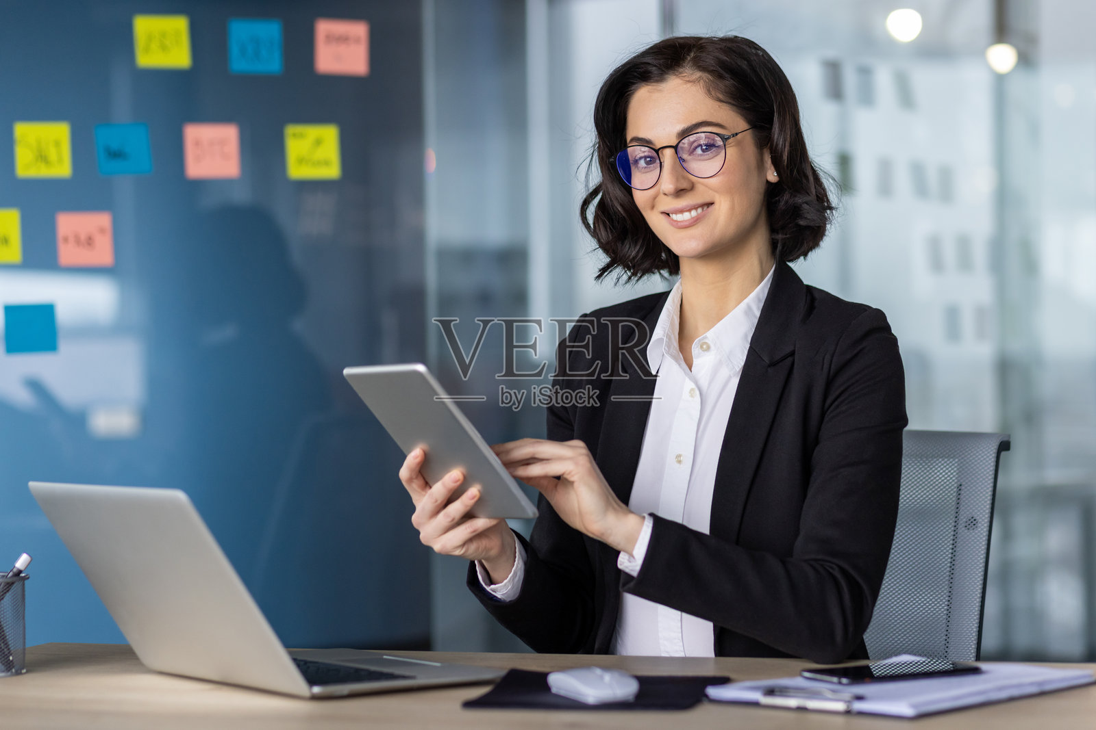 微笑的女商人在现代办公空间中使用平板电脑，同时处理笔记本电脑和文件。照片摄影图片