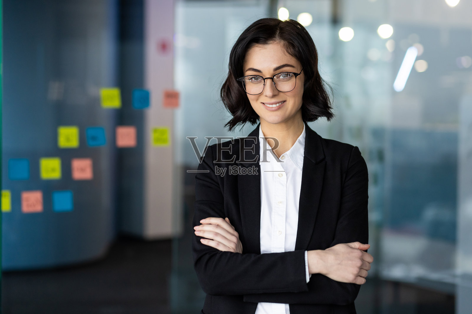 自信的年轻女商人在现代办公室里穿着黑色西装外套，戴着眼镜，双臂交叉，微笑着，周围是五颜六色的便利贴。照片摄影图片