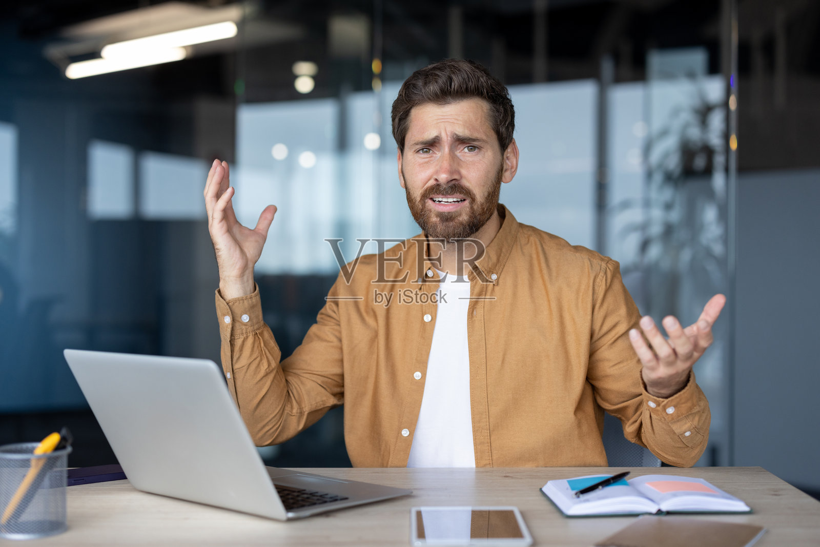 沮丧的商人在桌子前坐着，手势表达着对使用笔记本电脑的困惑，暗示着现代办公室环境中的技术问题，桌上还有笔记本和智能手机。照片摄影图片