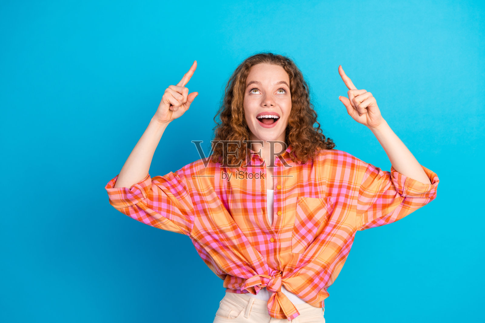 穿着格子衬衫的快乐年轻女性，卷发，指向明亮的蓝色背景。照片摄影图片