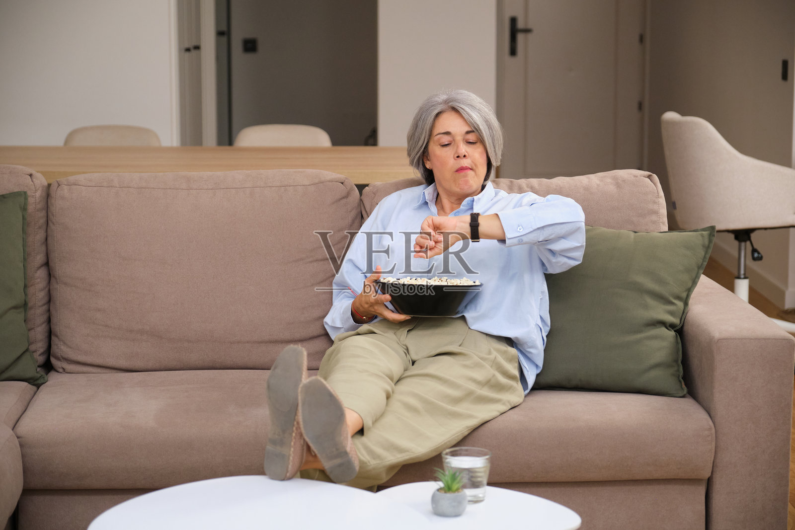 不耐烦的老年女性一边看电视一边吃爆米花，查看时间。照片摄影图片