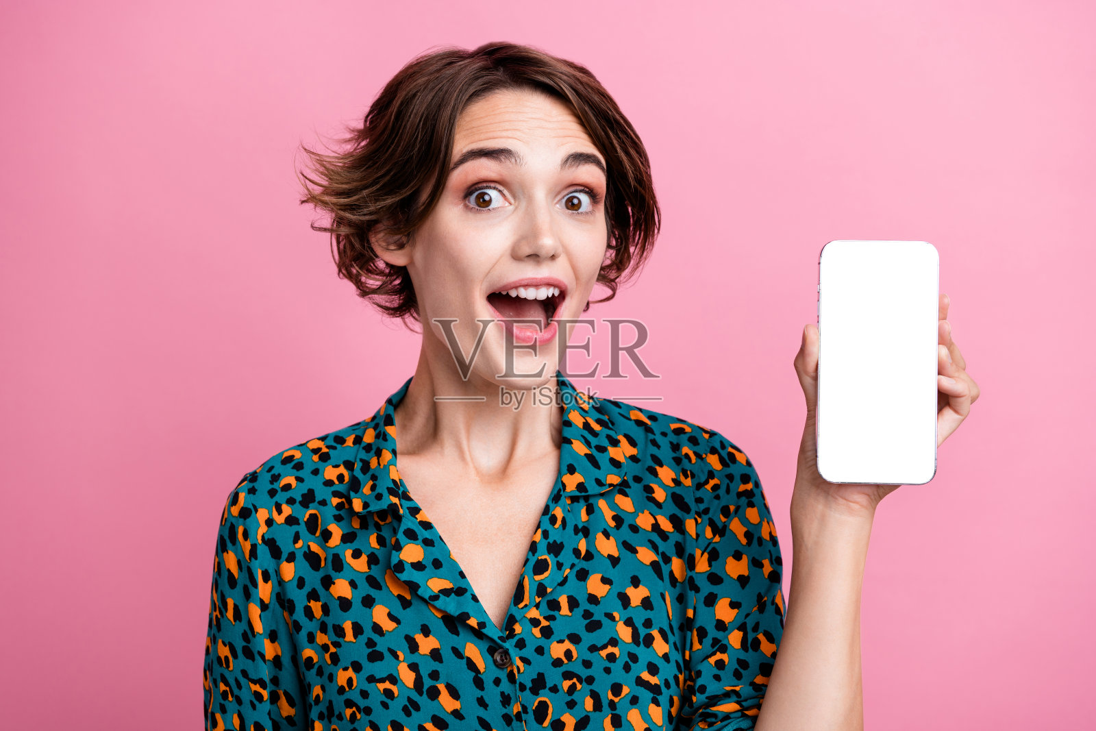 穿着印花衬衫的惊讶女性肖像，展示空白空间的电子商店，手持智能手机，背景为粉色。照片摄影图片