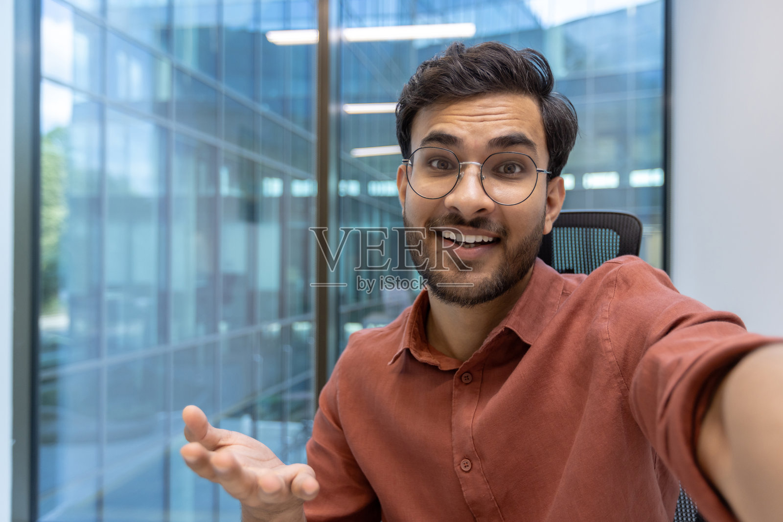 微笑的戴眼镜男子在现代办公室里用手机进行视频通话，正看着镜头。照片摄影图片