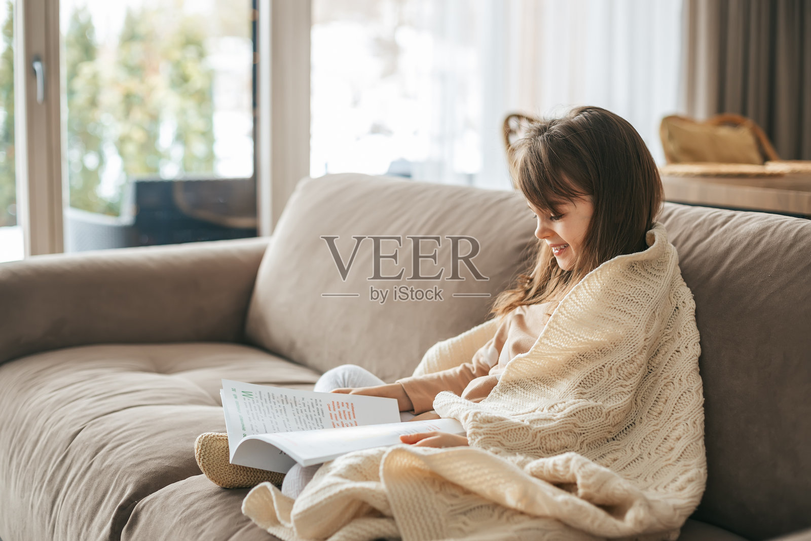 一个小女孩坐在沙发上，裹着一条奶油色的针织毛毯，手里拿着一本书。温馨的室内环境突显了安静阅读时刻的温暖与舒适。照片摄影图片