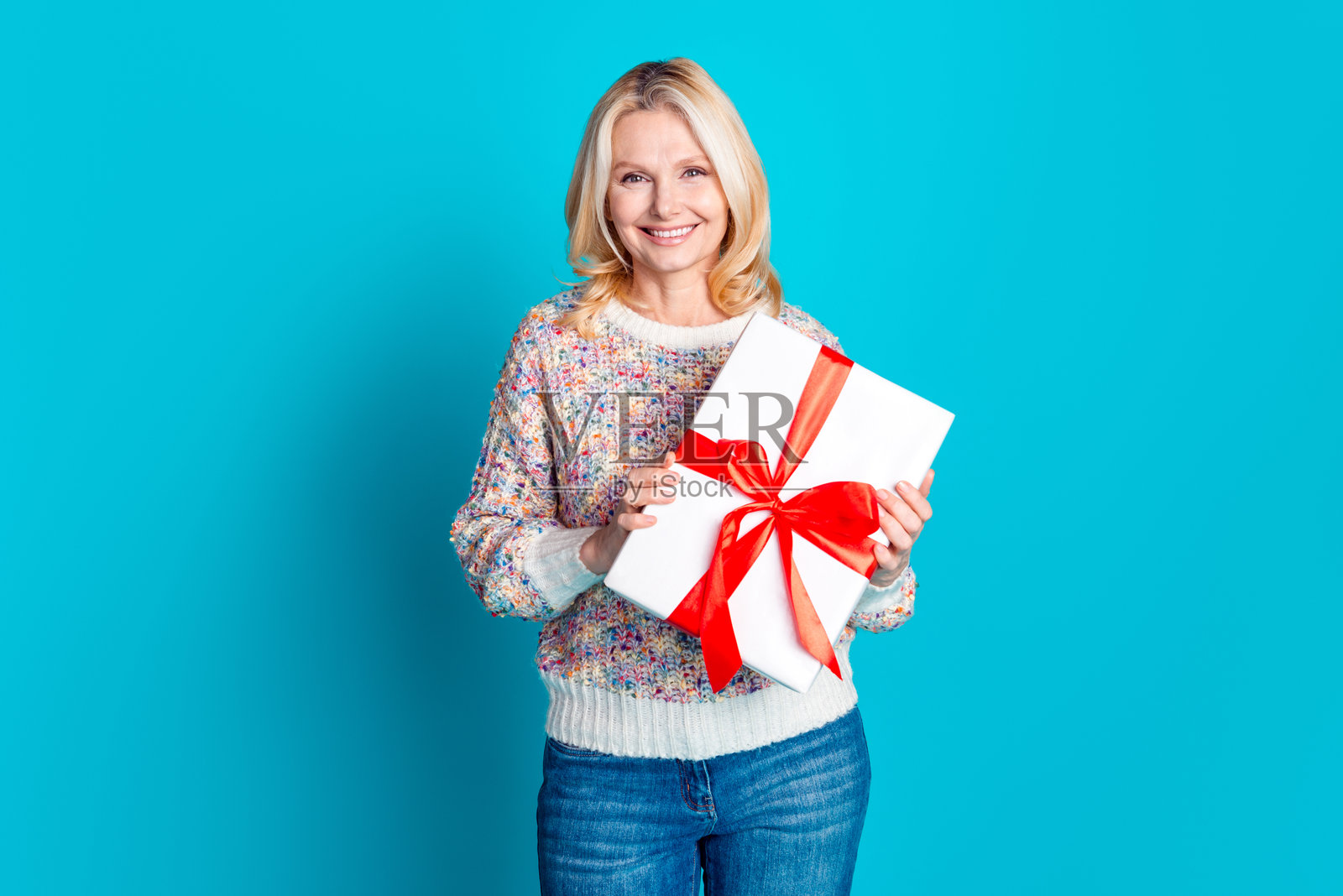 穿着毛衣的可爱老年女性手持礼品盒，背景为青绿色。照片摄影图片