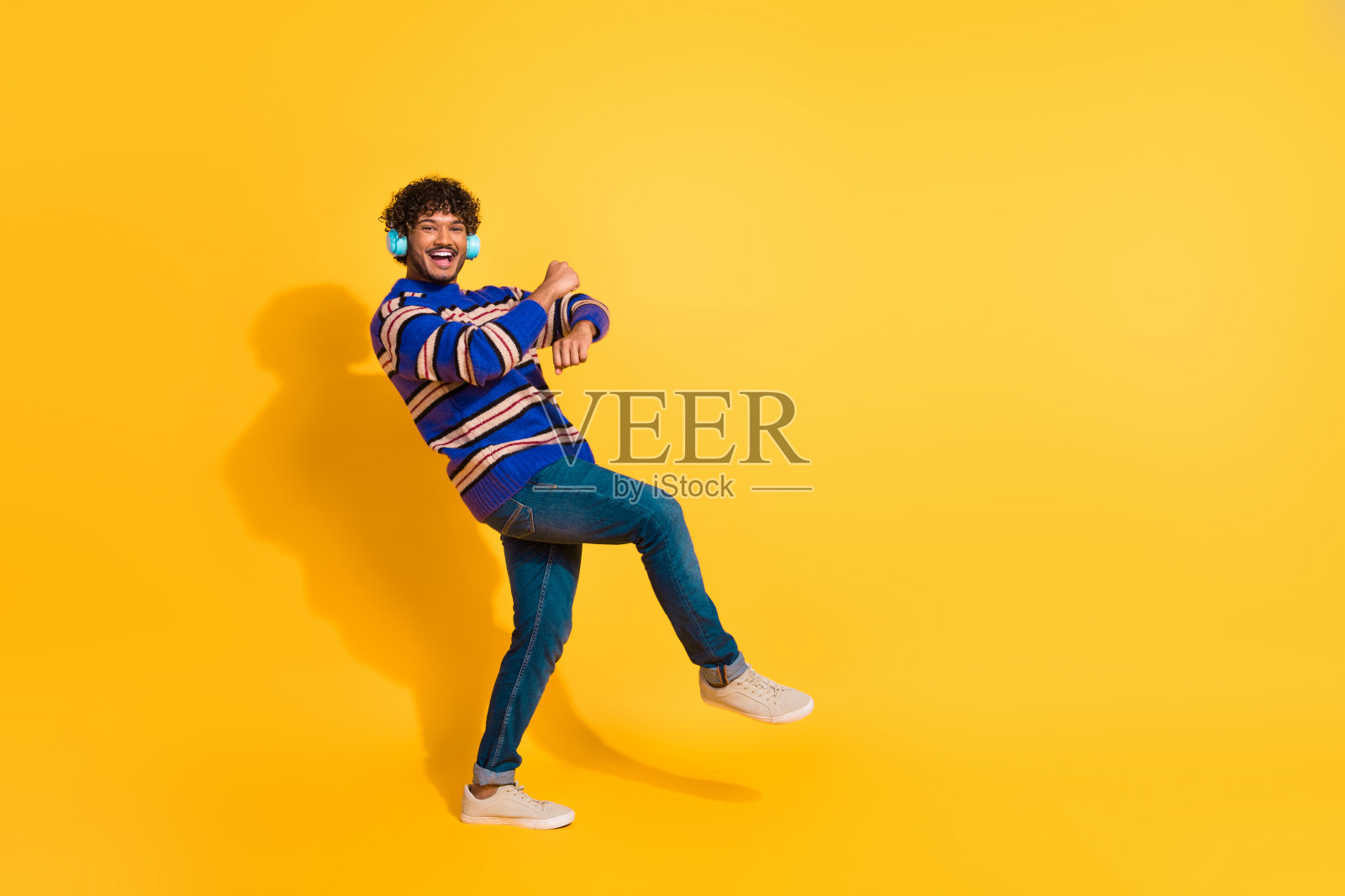 全身照，吸引人的年轻男子戴着耳机跳布吉舞，穿着蓝色条纹衣服，背景为黄色。照片摄影图片