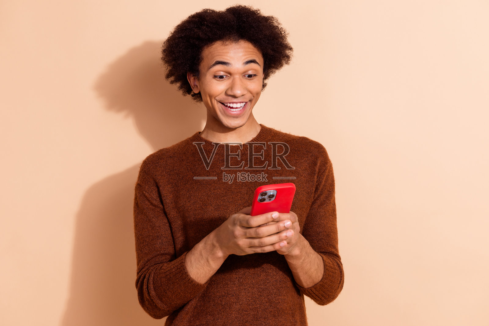 年轻男子兴奋地看着智能手机，发现在线促销，身穿休闲服装，站在米色背景前。照片摄影图片