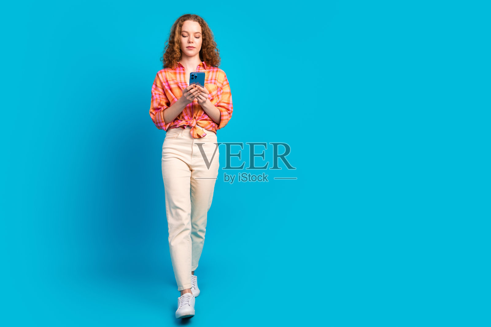 年轻的红发女性在明亮的蓝色背景下使用智能手机，传达出技术和连接感的休闲氛围照片摄影图片