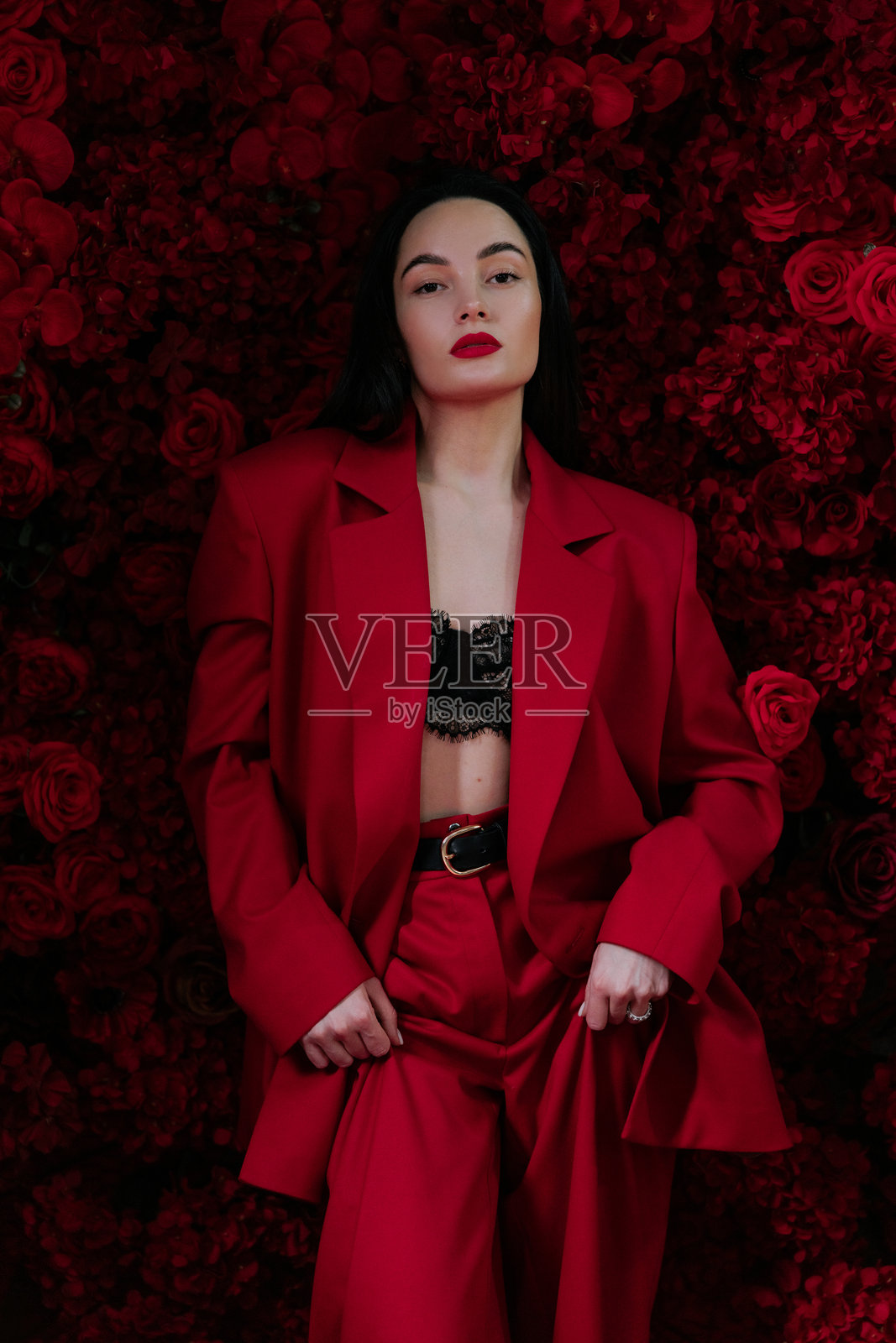 穿着红色裤子、夹克和黑色胸罩的美丽女孩在红玫瑰装饰的背景前摆姿势。照片摄影图片