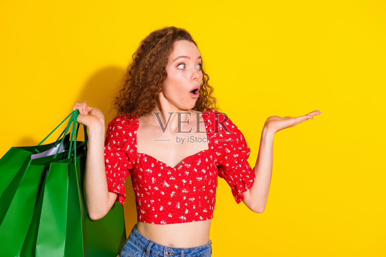 漂亮年轻女孩手持购物袋，面露惊恐，空旷的空间，穿着时尚的红色服装，背景为黄色。照片摄影图片