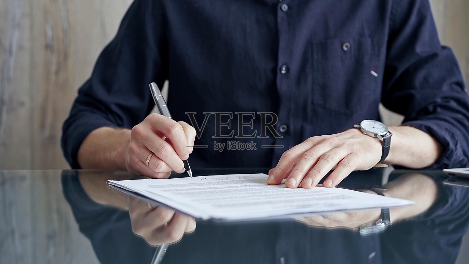 穿着深蓝色T恤的商人在桌子上签署合同。男性高管在玻璃桌上签署法律文件的特写。商业人士概念照片摄影图片