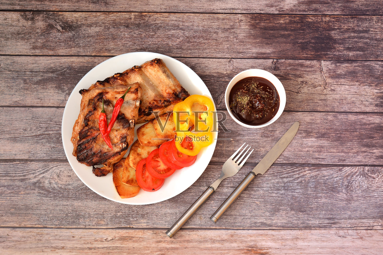 一盘烤猪肋排，配有土豆、甜椒和番茄，旁边有餐具和一杯酱汁。照片摄影图片