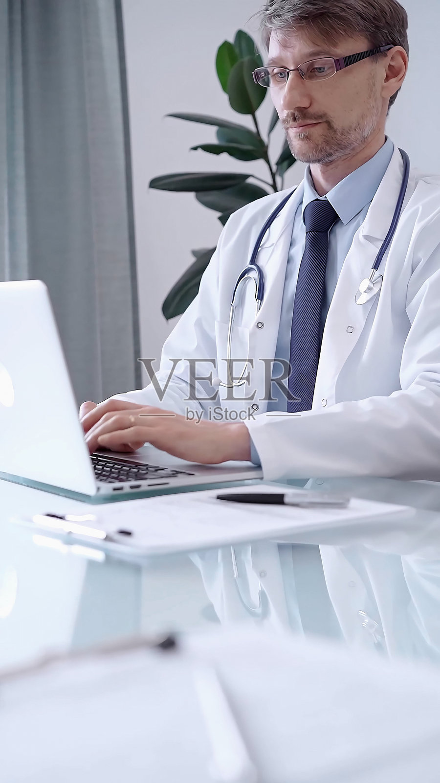 身穿白大褂的男性医生专注于现代诊所办公室的笔记本电脑数据，同时分析患者信息。医学概念照片摄影图片