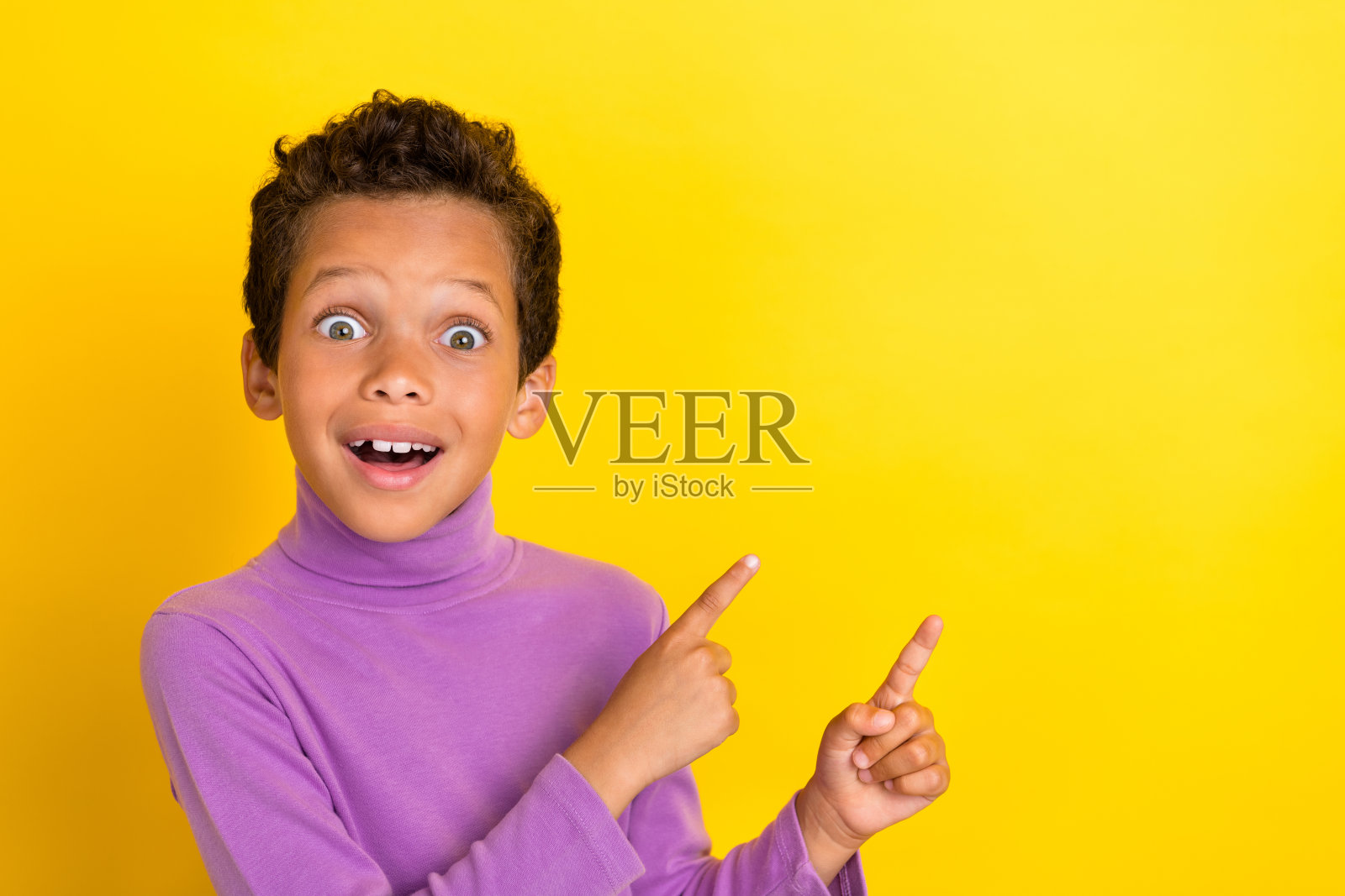 可爱的男孩惊讶地指着空白处，难以置信的折扣，穿着时尚的紫色服装，背景为黄色。照片摄影图片