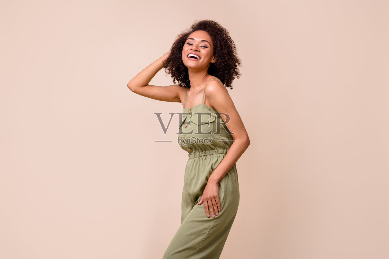 美丽年轻女性触摸波浪形发型，展示新造型，穿着时尚的卡其色服装，背景为米色。照片摄影图片