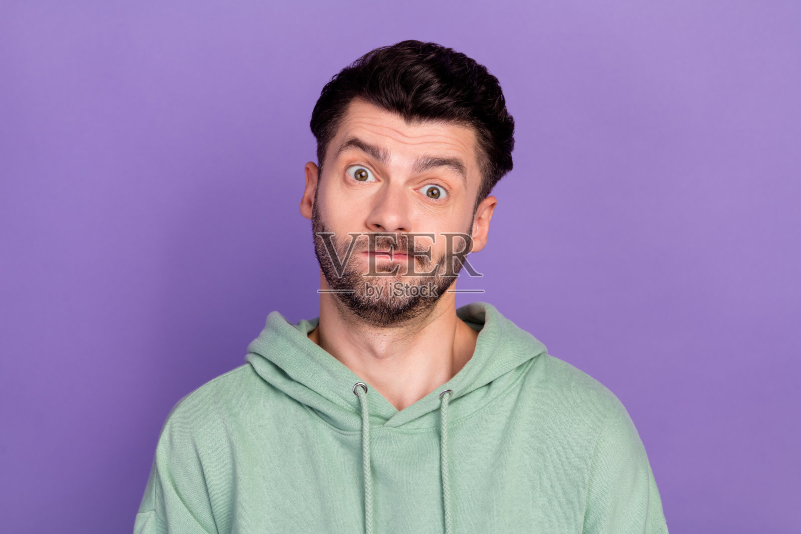 年轻有趣的不开心男士特写，穿着卡其色连帽衫，撅着嘴唇，对自己新发型感到不满意，背景为紫色。照片摄影图片