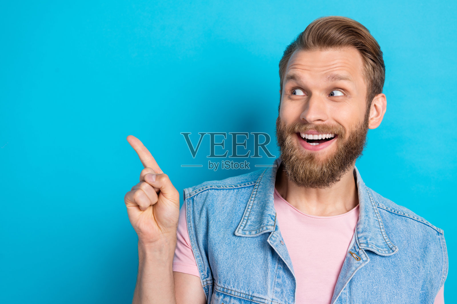 穿着牛仔背心的兴奋年轻男子，手指指向空旷的空间，背景为蓝色。照片摄影图片