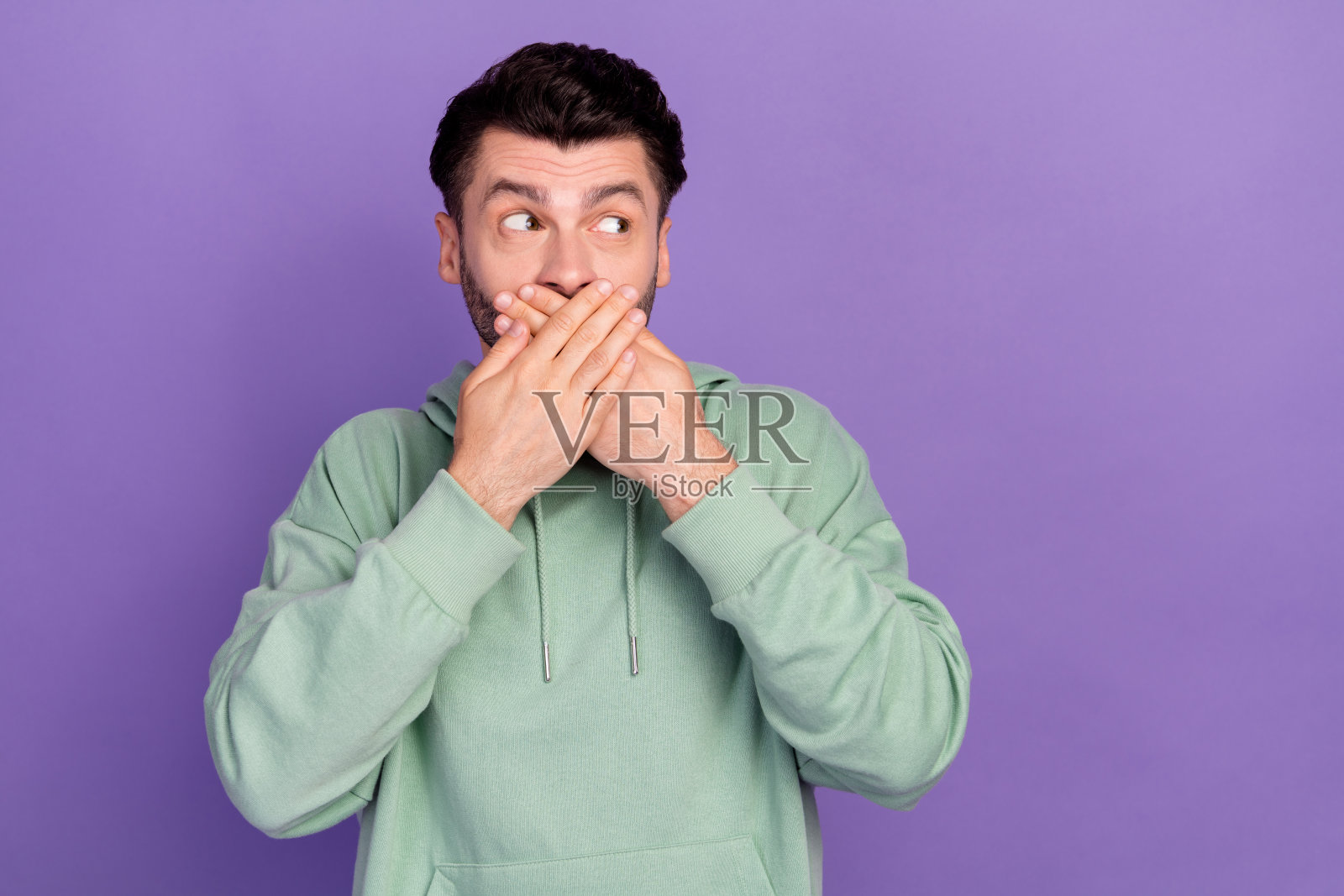 年轻兴奋的积极男性肖像，双手捂嘴安静地笑，目光投向空旷的地方，似乎在分享有趣的秘密，背景为紫色。照片摄影图片
