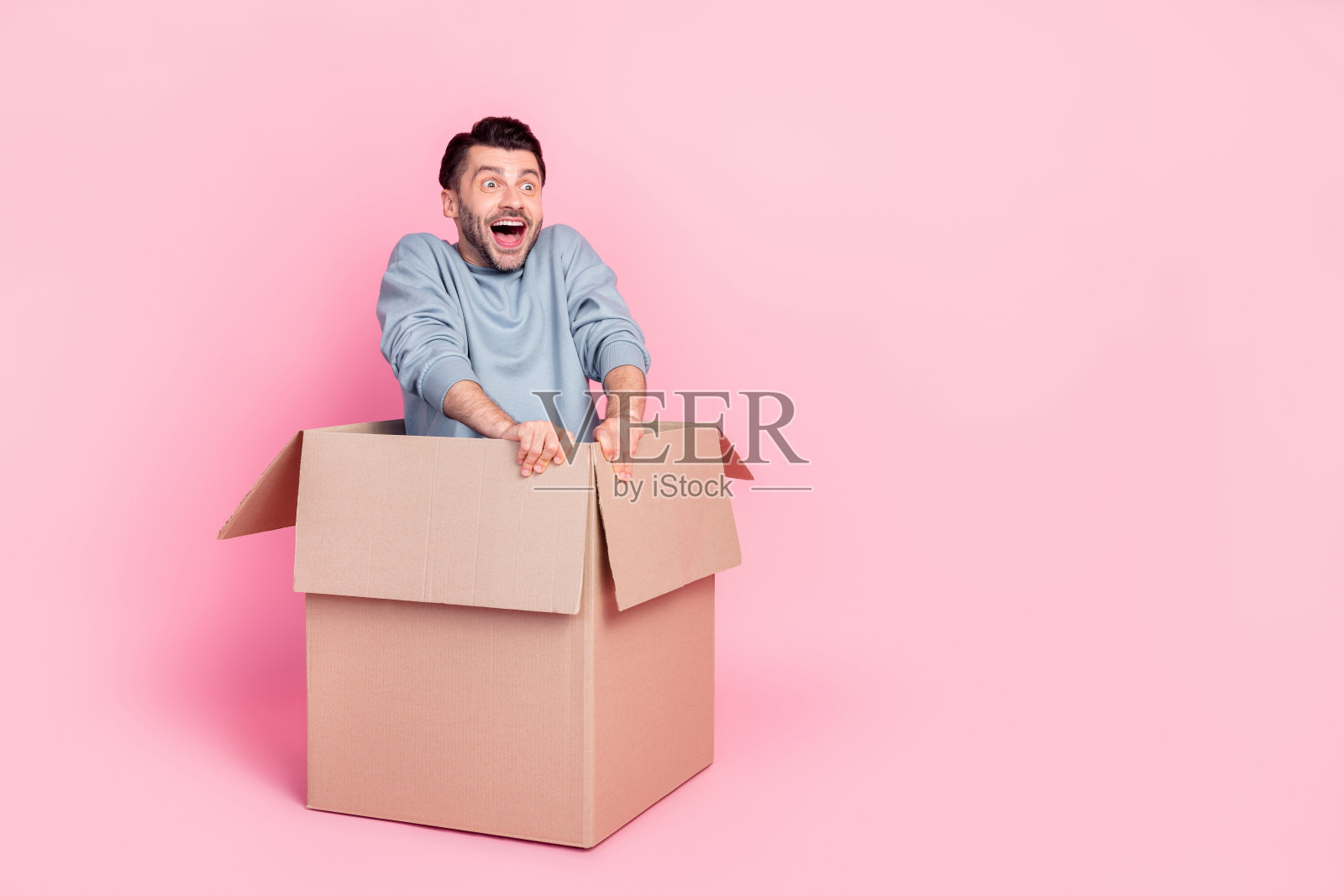 全身照，年轻帅气的男孩兴奋地坐在箱子上，准备搬家，穿着时尚的蓝色服装，背景为粉色。照片摄影图片