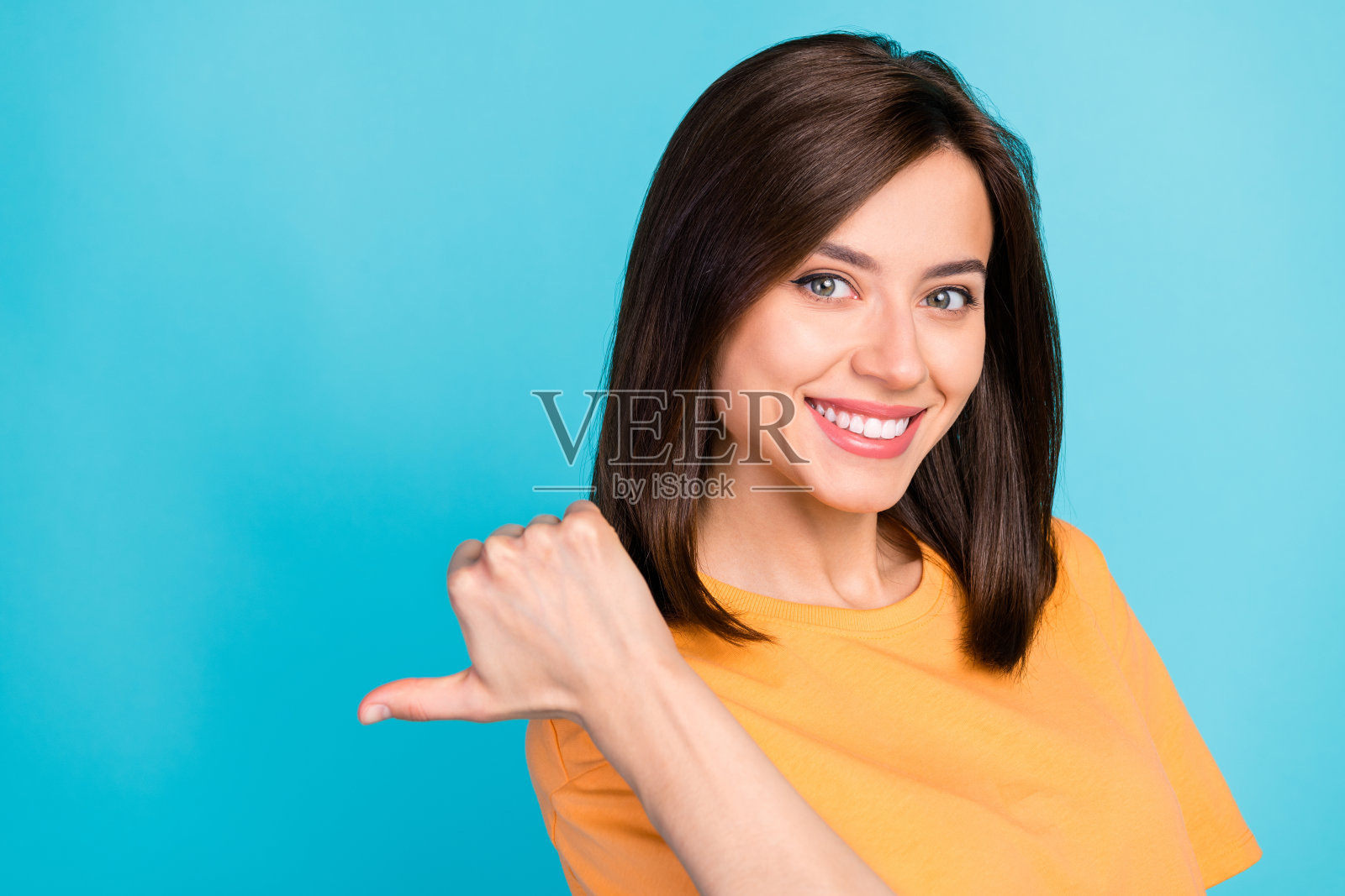 一位露齿微笑、愉快的女孩，直发，穿着黄色T恤，指向蓝色背景中的空白处。照片摄影图片