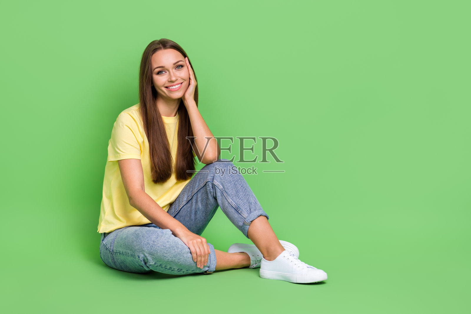穿着时尚黄色衣服、牛仔裤和运动鞋的积极酷女孩坐在地板上，背景为绿色。照片摄影图片