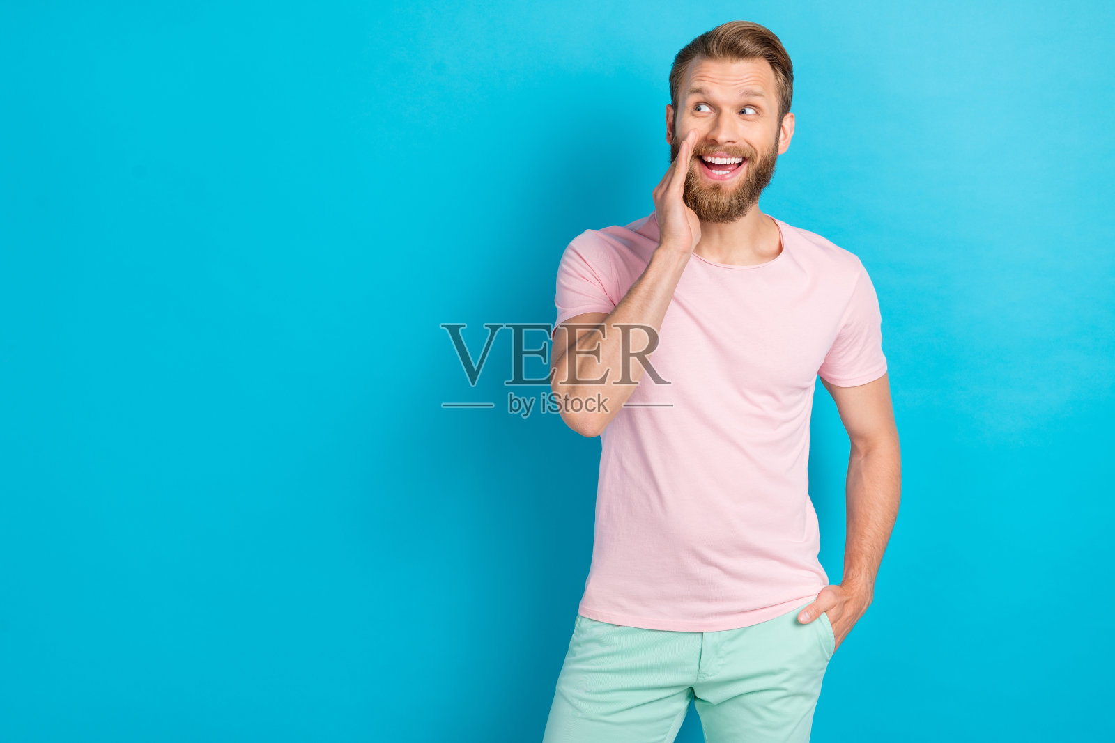 穿着粉色T恤的时尚男子，惊讶地用手捂住嘴巴，似乎在对着空旷的蓝色背景讲秘密。照片摄影图片