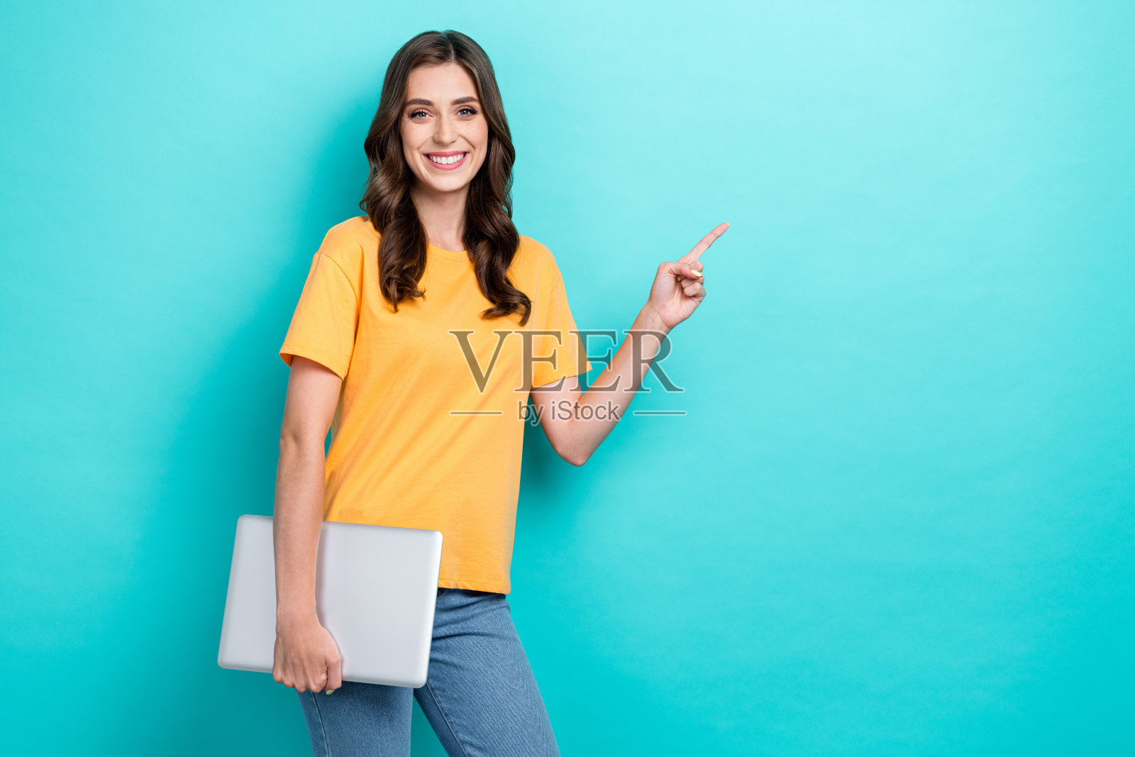 年轻漂亮的可爱女性穿着黄色T恤，手指指向空白处，手持电脑推荐电子购物，背景为青色。照片摄影图片
